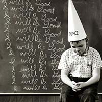 photo of a young male sitting on a stool with a dunce cap on in front of a chalkboard with I will be good written on it many times