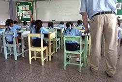 photo of several students sitting at desks taking a test while a male teacher stands watching over them